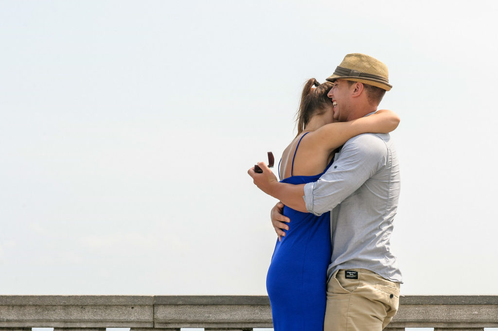 Man hugs his fiancée while holding ring box