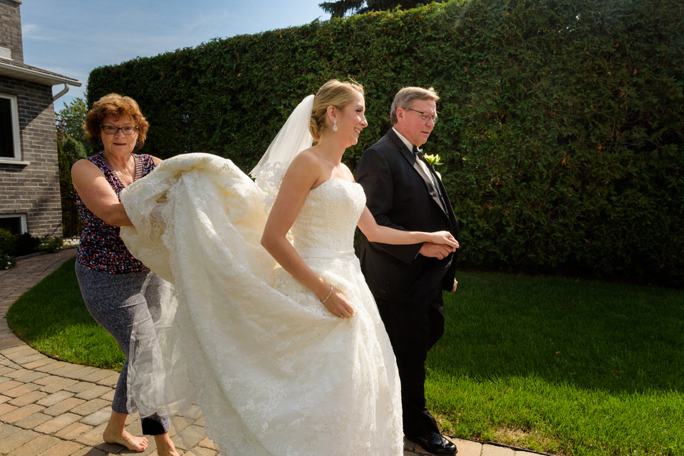 Bride leaving her house with her dad and a neighbour helping with her dress