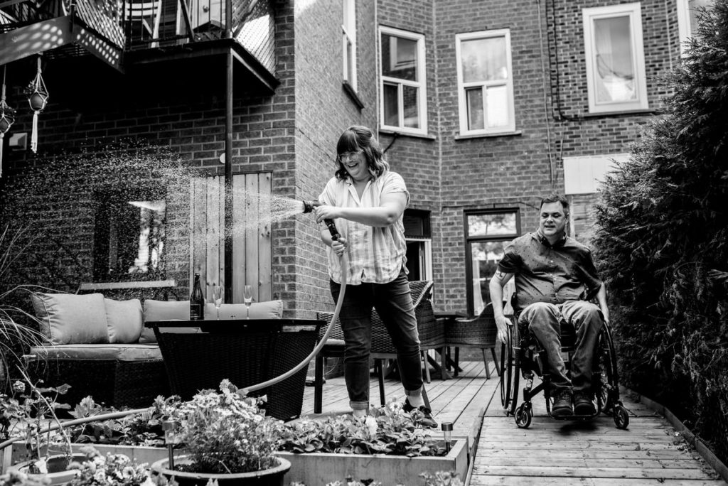 Woman waters flowers in backyard garden while man heads down wheelchair ramp